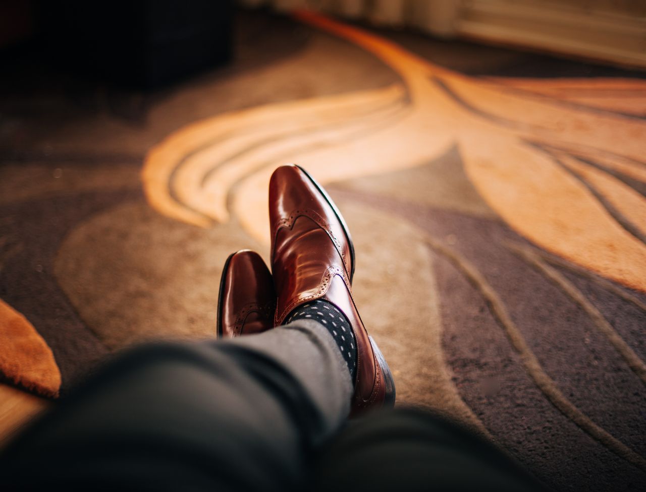 collection-01 Close-up of polished brown leather shoes worn with patterned socks, relaxed indoors.