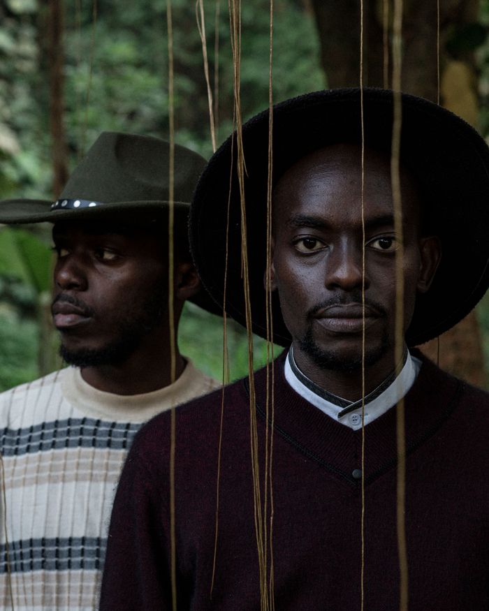our-mission-01 Two men wearing stylish hats, standing amidst greenery, captured in a moody outdoor setting.
