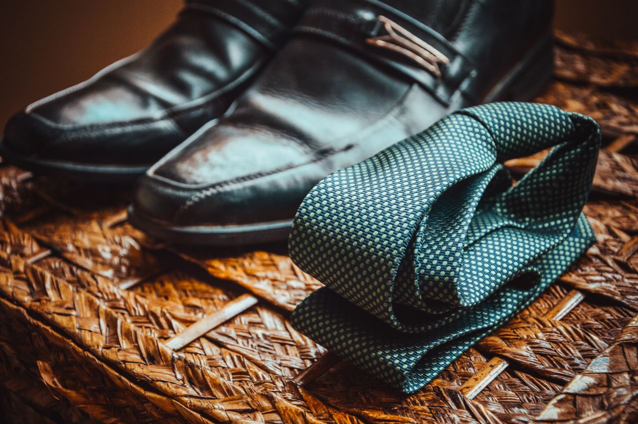 our-story-02 Close-up of a stylish necktie and polished leather shoes resting on a woven wicker surface.